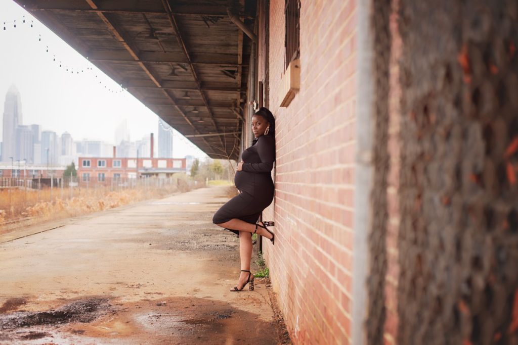 pregnant woman leaning on brick wall charlotte skyline in background urban warehouse
