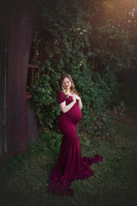 pregnant woman touching chest in front of rusty barn and overgrown foliage